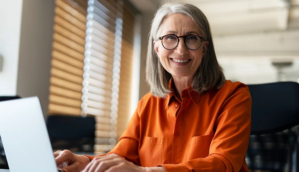 Professional senior woman with glasses and grey hair smiling while working on a laptop.